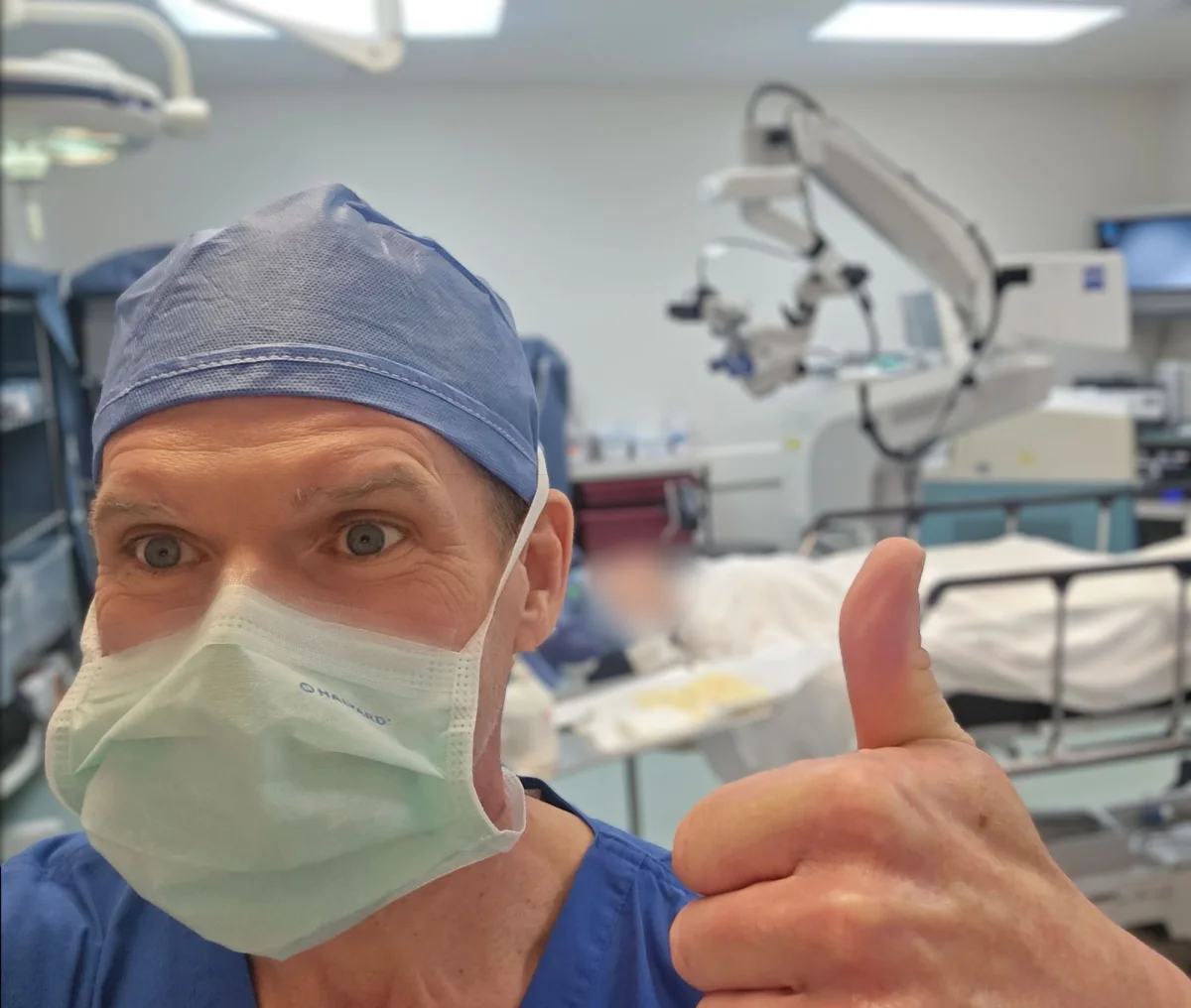 Marc Jones in scrubs, cap and surgical mask, giving a thumbs-up in the operating room